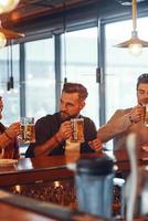 Handsome young men in casual clothing enjoying beer while sitting at the bar counter in pub photo