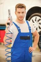 Confident expert. Confident young man holding a pneumatic wrench and looking at camera while standing in workshop with car in the background photo