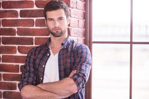 Charming and confident. Handsome young man keeping arms crossed and looking at camera while standing indoors photo