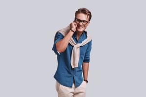 Good talk. Handsome young man talking on the smart phone and looking away with a smile while standing against grey background photo