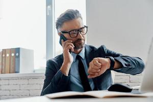 Handsome mature man in formalwear checking the time while talking on mobile phone in the office photo