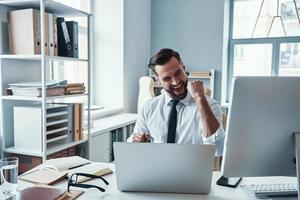 alegre joven con camisa y corbata trabajando usando computadora y sonriendo mientras está sentado en la oficina foto