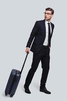 In a hurry. Full length of good looking young man with suitcase looking away while standing against grey background photo