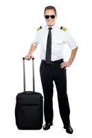 New day and new destination. Confident male pilot in uniform leaning hand on his suitcase and smiling while being isolated on white background photo