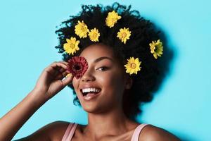 Natural beauty. Attractive young African woman looking at camera and covering eye with a flower while lying against blue background photo