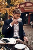 Enjoying fresh coffee. Handsome young man in smart casual wear drinking coffee while sitting in restaurant outdoors photo