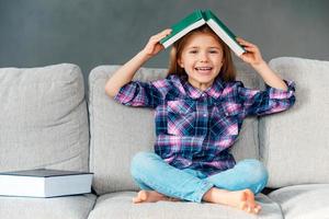 Knowledge assimilate better this way  Cheerful little girl holding book over her head and looking at camera with smile while sitting on the couch in lotus position at home photo