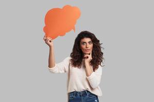 This is an idea Thoughtful young woman staring at camera and touching her face with index finger while standing against grey background photo