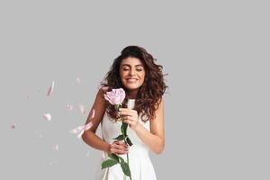 mujer joven feliz sonriendo y sosteniendo una flor mientras está de pie contra un fondo gris con pétalos de rosa volando alrededor foto