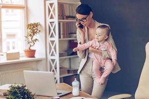 no es fácil ser una madre trabajadora joven y hermosa mujer de negocios hablando por teléfono móvil y mirando una computadora portátil mientras está de pie con su bebé en su lugar de trabajo foto