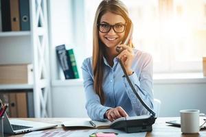 I will connect you in one second Cheerful young beautiful woman in glasses talking on the phone and looking at camera with smile while sitting at her working place photo