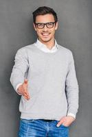 Nice to meet you Cheerful young man reaching out his hand in greeting you and holding hand in pocket while standing against grey background photo