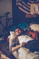 Young handsome man looking away while lying down on sofa in messy room after party photo