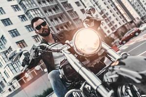 Dangerous. Handsome young man in leather jacket and sunglasses riding motorbike while spending time outdoors photo