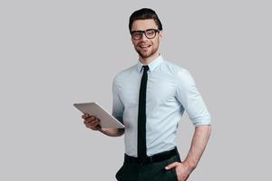 experto en negocios confiado. un joven apuesto con camisa blanca y corbata sosteniendo una tableta digital y sonriendo mientras se enfrenta a un fondo gris foto