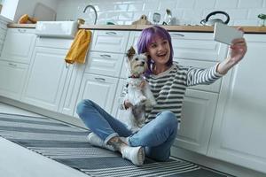 mujer feliz cargando un perrito y haciéndose selfie mientras se sienta en el suelo en la cocina foto