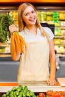 vitaminas para los clientes. alegre joven vendedora sonriendo a la cámara y sosteniendo zanahorias mientras está de pie en una tienda de alimentos foto