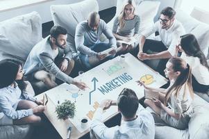 Discussing strategy together. Top view of business people discussing something while sitting around the desk together and pointing large paper with graphs and charts photo