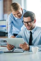 Discussing new business project. Two happy young people in formalwear smiling and looking at digital tablet while working together photo