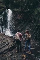 en el maravilloso lugar. vista trasera completa de una pareja joven tomándose de la mano mientras está de pie cerca de la cascada foto