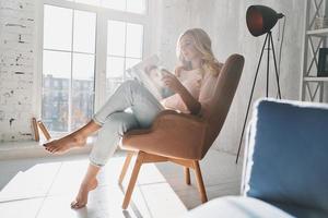 al tanto de las últimas noticias de moda. hermosa joven leyendo una revista mientras se sienta en el sillón en casa foto