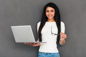 How may I help you Attractive young African woman holding laptop and looking at camera with smile while standing against grey background photo