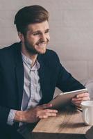 Great news Handsome young man holding his digital tablet and looking away with smile while sitting at his working place photo