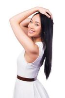 Feeling flirty. Side view of beautiful young Asian woman in pretty dress keeping hands in hair and smiling while standing against white background photo