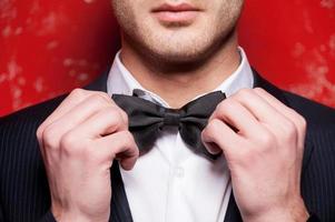 Tying his bow tie. Cropped image of handsome young man in formalwear adjusting his bow tie while standing against red background photo