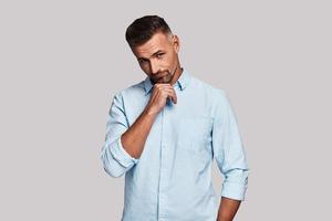 Taking time to think. Thoughtful young man keeping hand on chin and making a face while standing against grey background photo