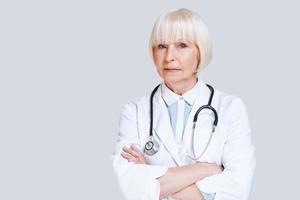 General practitioner. Beautiful senior woman in lab coat looking at camera and keeping arms crossed while standing against grey background photo