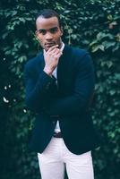 Thoughtful handsome. Handsome young African man in smart casual wear holding hand on chin and looking at camera while standing against green plant background outdoors photo