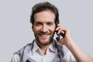 Pleasant talk. Beautiful young man in smart casual clothes talking on the phone and looking at camera while standing against grey background photo