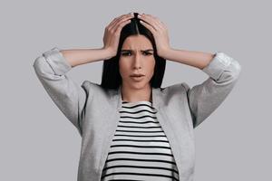 Terrible headache. Frustrated young woman in smart casual wear touching her head with hands and making face while standing against grey background photo