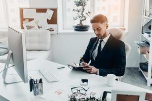 Making some notes. Handsome young man in full suit writing something in his notebook while sitting at the office desk photo
