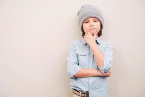 Deep in his thoughts. Handsome little boy in hat holding hand on chin and looking at camera while standing against grey background photo