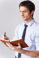 escribiendo sus pensamientos. un joven confiado con camisa y corbata escribiendo algo en un bloc de notas mientras se enfrenta a un fondo gris foto