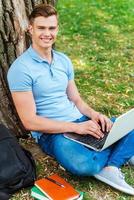 Finding a peaceful place for studying. Top view of confident male student working on laptop and smiling while sitting on the grass and leaning at the tree photo