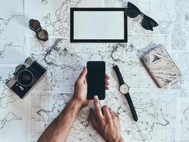 Keep traveling. Close up top view of man using smart phone with sunglasses, photo camera, compass, watch and passport lying on map around