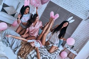 Nacido para divertirse, mujeres jóvenes juguetonas tratando de ponerse orejas de conejo en el pie de sus novias y sonriendo mientras se sientan en la cama. foto