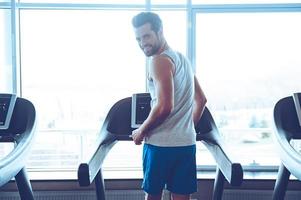 Enjoying his exercising. Rear view of young handsome man in sportswear running on treadmill in front of window at gym and looking at camera with smile photo