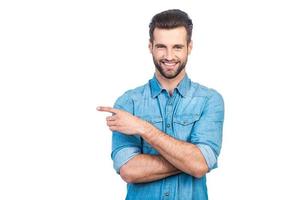 mire hacia allí un joven apuesto feliz con camisa de jeans apuntando hacia otro lado y sonriendo mientras está de pie contra el fondo blanco foto