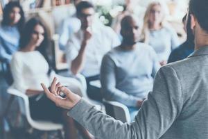 Business coach. Rear view of man gesturing with hand while standing against defocused group of people sitting at the chairs in front of him photo