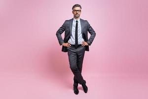 Confident mature man in full suit holding hands on hip while standing against pink background photo