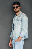 Cool and trendy. Side view of confident young man in sunglasses holding hand in pocket and looking at camera while standing against grey background photo