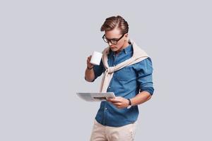 Reading latest news.  Handsome young man reading newspaper and holding a cup while standing against grey background photo