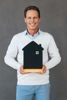 Ready to move in. Confident mature man holding house shaped object in hand and smiling while standing against grey background photo
