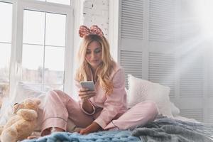 Just relaxing. Beautiful young smiling woman using her smart phone while sitting on the bed at home photo