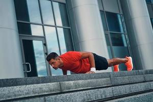Athletic young man in sports clothing keeping plank position while exercising outdoors photo
