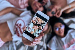 autofoto perfecto vista superior de cuatro mujeres sonrientes jóvenes y juguetonas con anteojos tomándose selfie con un teléfono inteligente mientras están acostadas en la cama en casa foto